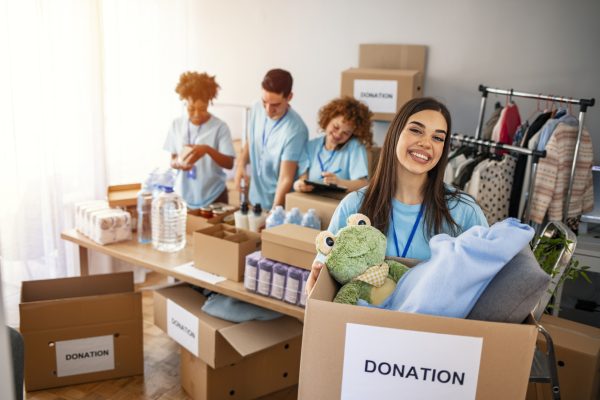 We are successful team of social workers. Woman volunteer holding a donation box and is smiling at the camera. Volunteers are helping people in the background.